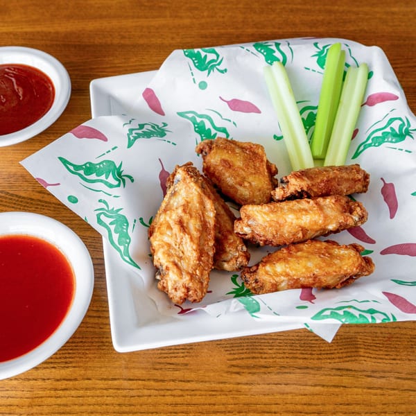 Crispy fried chicken wings with celery sticks and two bowls of red sauce on a wooden table.