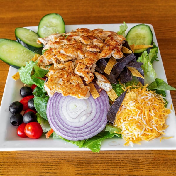 A vibrant Mexican chicken salad with lettuce, red onion, tomatoes, olives, cucumbers, cheese, and tortilla strips.
