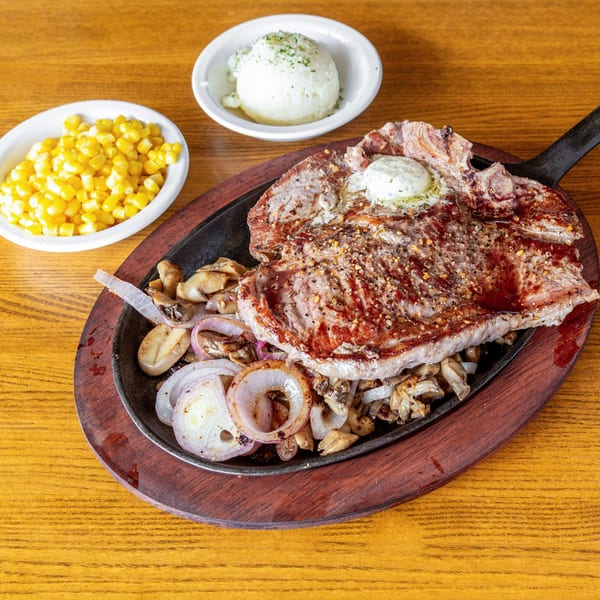 Sizzling steak with butter, onions, mushrooms, corn, and mashed potatoes on a wooden table.